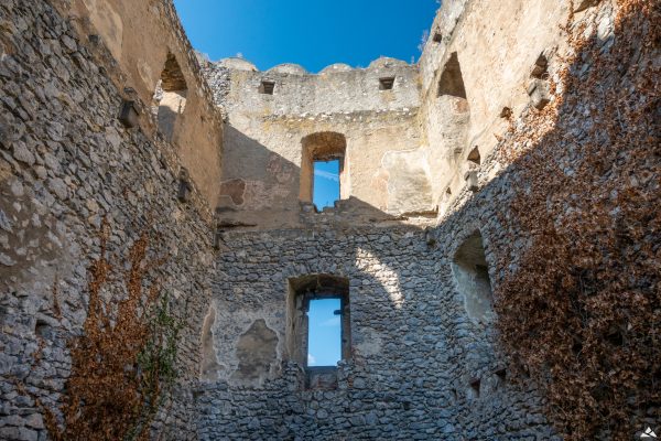 Kadr wypełniony przez kamienne mury. Na trzech ścianach (lewej, środkowej i prawej), znajduje się 5 prostokątnych otworów okiennych. Część ścian prawej i lewej porośnięta jest przez bluszcze.