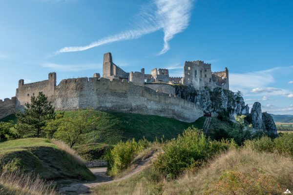 W centralnej części ruiny zamku Beckov - wysokie, kamienne mury obronne; kaplica zamkowa oraz wieże obronne. Na pierwszym planie: zarośla oraz drzewa.