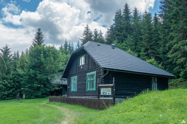 Ciemnobrązowy, drewniany budynek z dwoma zielonymi oknami. Wokół zielona łąka i las.